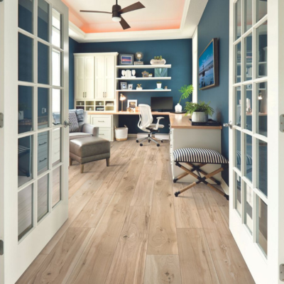 Home office with light wood laminate flooring, deep blue walls, and an L-shaped desk featuring built-in storage, a white office chair, and decorative plants.
